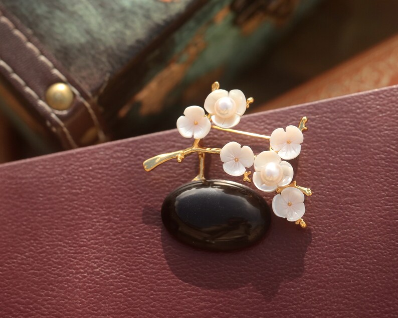 May include: A gold-toned brooch featuring a black oval base with a delicate branch of white flower blossoms. Each blossom has a pearl center. The brooch is set against a burgundy leather surface.