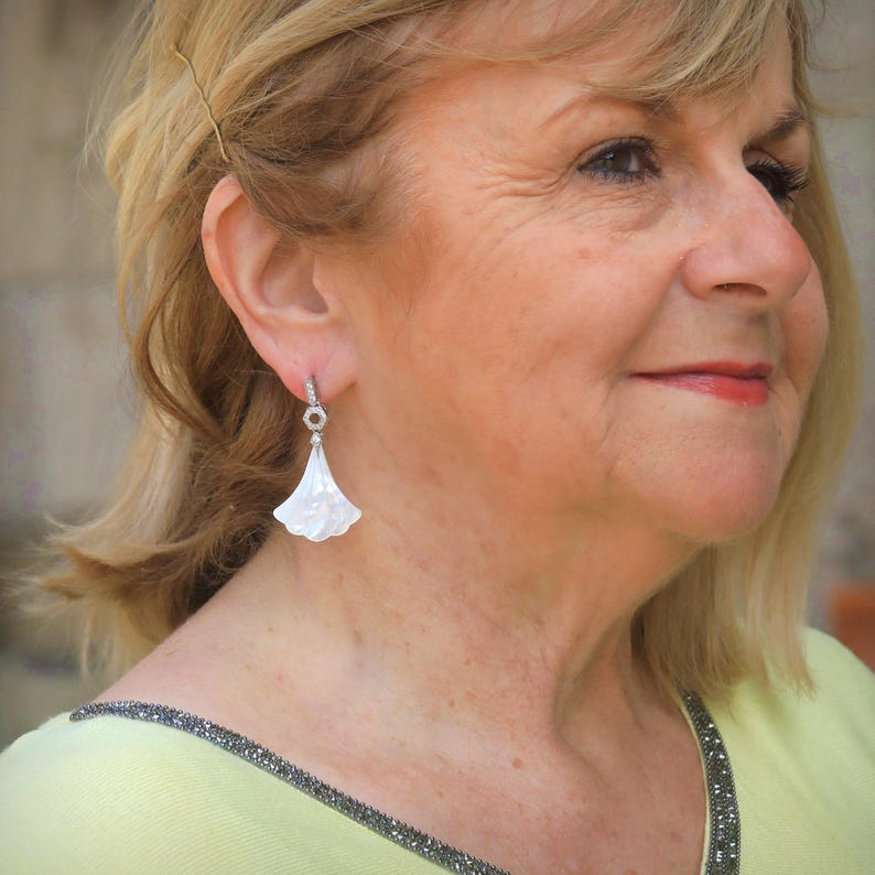 May include: A close-up of a woman wearing a pair of white, fan-shaped earrings. The earrings have a small, circular top with a clear stone and a white, petal-like design that hangs down. The woman is wearing a light yellow sweater.