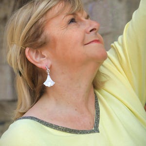 May include: A woman wearing a light yellow top with a beaded neckline and a pair of white, fan-shaped earrings. The earrings are dangling from her ears. The woman is looking up, with a soft focus on her face and the earrings.