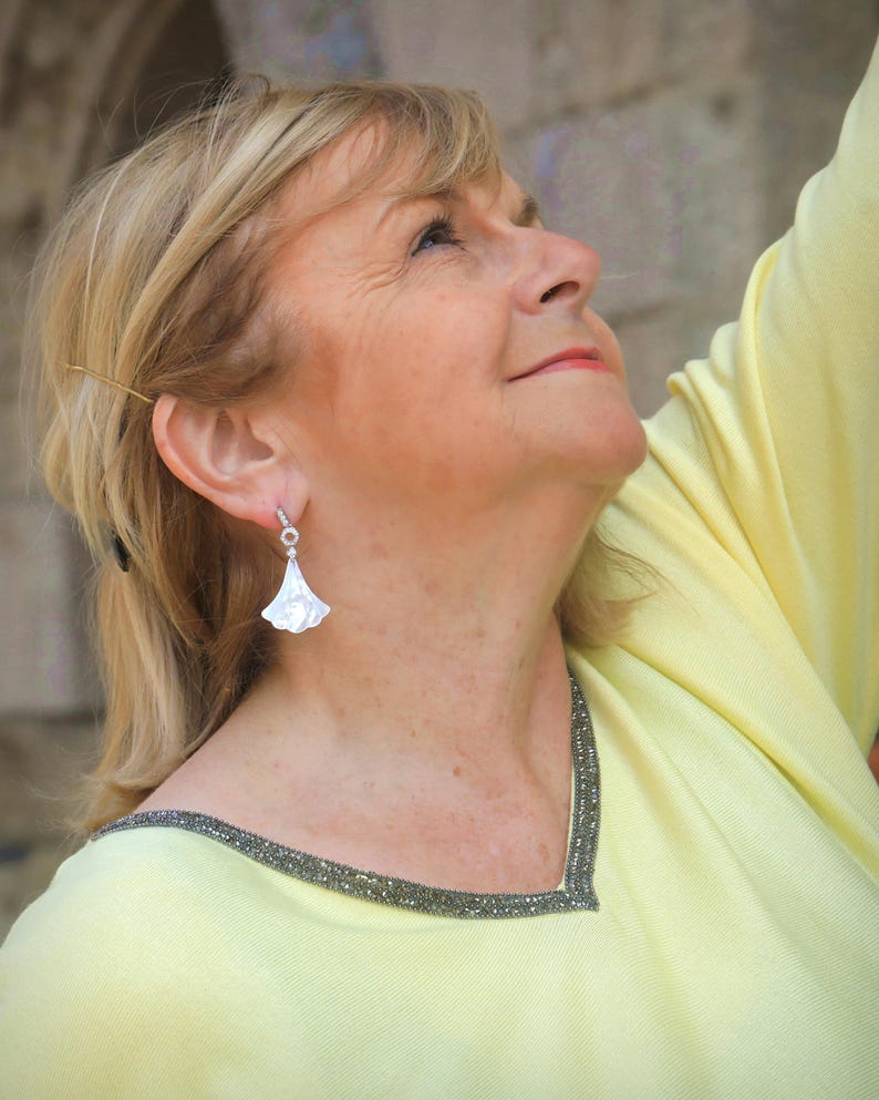 May include: A woman wearing a light yellow top with a beaded neckline and a pair of white, fan-shaped earrings. The earrings are dangling from her ears. The woman is looking up, with a soft focus on her face and the earrings.