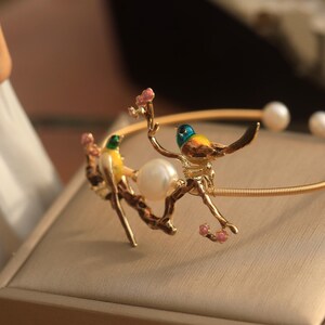 May include: A gold bracelet with two yellow and green birds perched on a branch with pink flowers. The bracelet has a pearl accent.