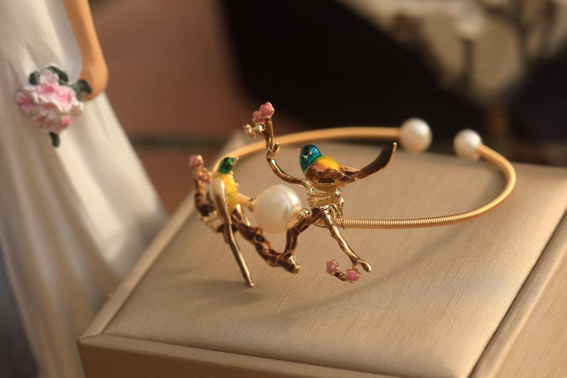 May include: A gold bracelet with two yellow and green birds perched on a branch with pink flowers. The bracelet has a pearl accent.