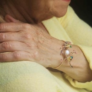May include: A gold bangle bracelet with a delicate design featuring a pearl and colorful enamel accents. The bracelet is adorned with small flowers and birds.