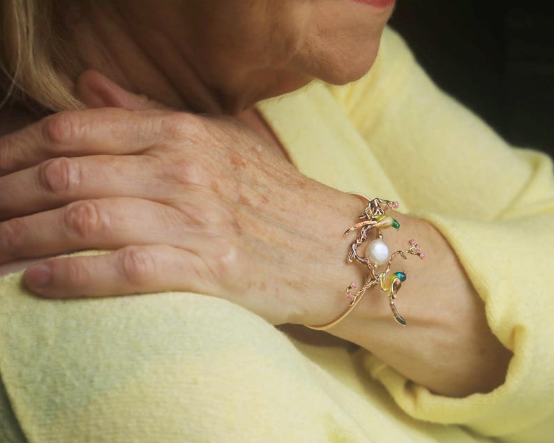 May include: A gold bangle bracelet with a delicate design featuring a pearl and colorful enamel accents. The bracelet is adorned with small flowers and birds.