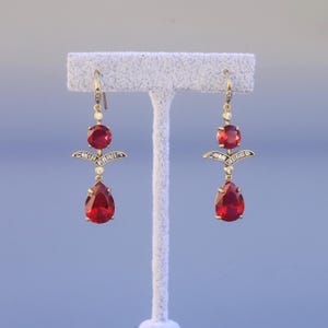 Könnte beinhalten: Ein Paar goldfarbene Ohrhänger mit roten Edelsteinakzenten. Jeder Ohrring hat einen runden roten Stein oben, ein dekoratives Goldelement mit kleinen weißen Steinen und einen tropfenförmigen roten Stein unten. Die Ohrringe werden auf einem weißen Ständer präsentiert.