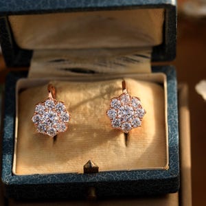Puede incluir: Un par de pendientes de oro rosa con un racimo de piedras de circonita c&uacute;bica transparentes en forma de flor. Los pendientes est&aacute;n en una peque&ntilde;a caja de terciopelo.