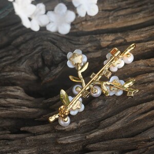 May include: A gold-toned brooch in the shape of a delicate floral branch with white pearl-like flowers and leaves. The brooch is on a dark brown wooden surface.