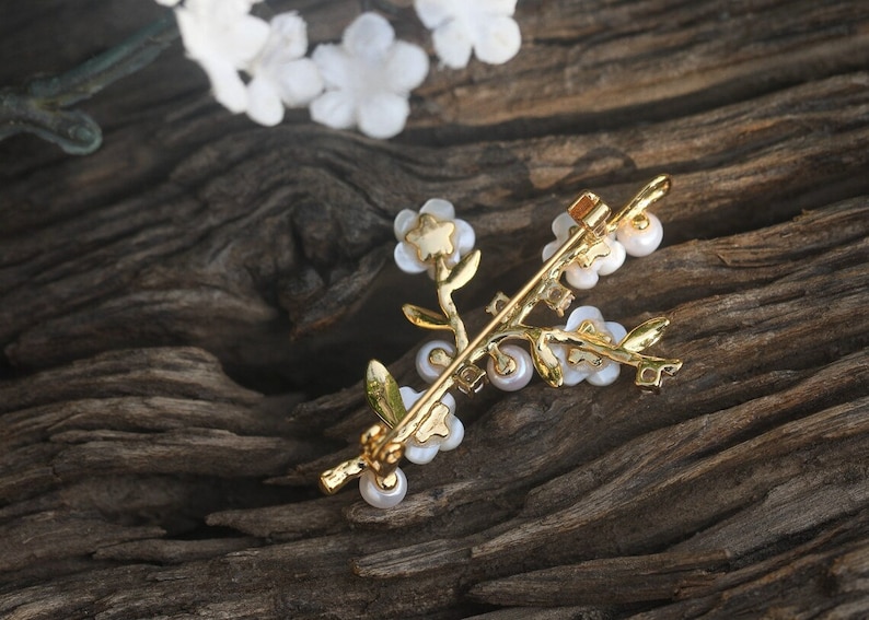 May include: A gold-toned brooch in the shape of a delicate floral branch with white pearl-like flowers and leaves. The brooch is on a dark brown wooden surface.
