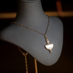 May include: A gold-toned necklace featuring a geometric pendant with a central white mother-of-pearl semi-circle and a small clear round stone set above it. The pendant has vertical metallic bars and hangs from a delicate gold-toned chain.