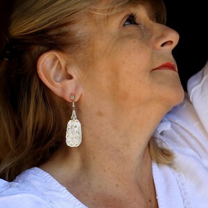 May include: A woman wearing a white blouse with a delicate floral pattern, with a pair of silver earrings featuring a carved, pearlescent design.