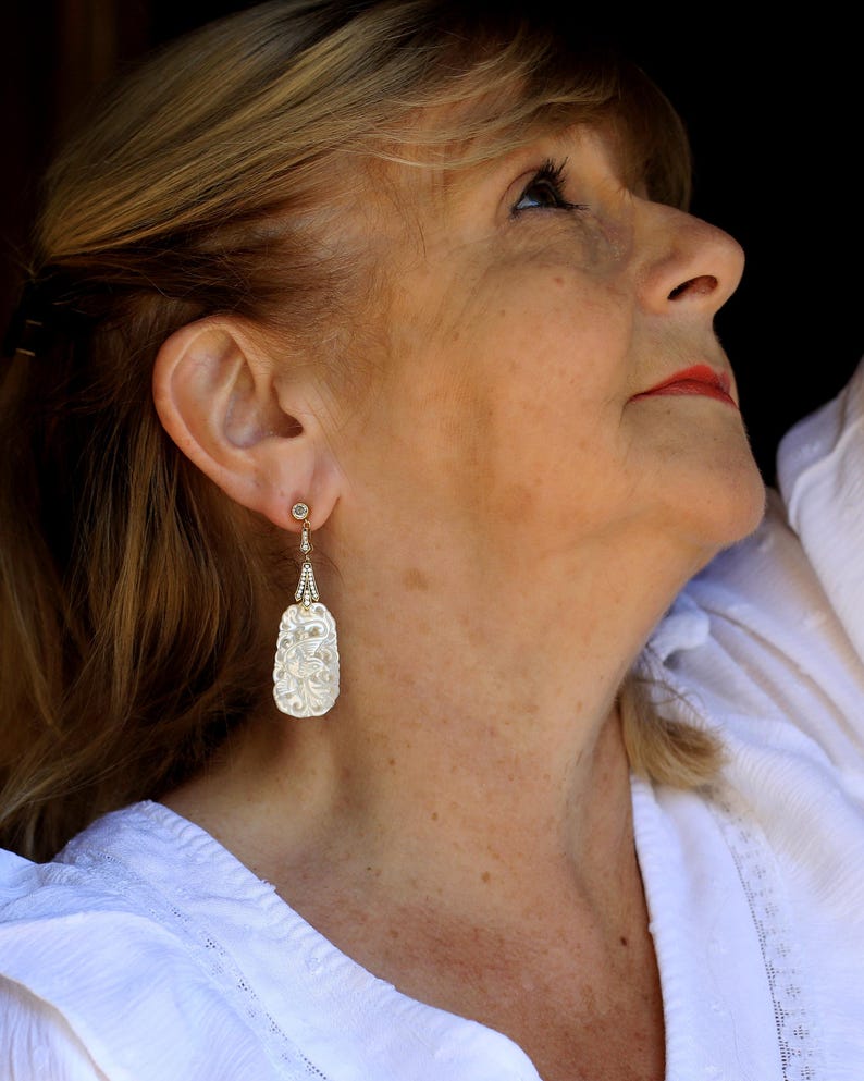 May include: A woman wearing a white blouse with a delicate floral pattern, with a pair of silver earrings featuring a carved, pearlescent design.