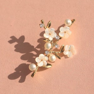 May include: Gold tone brooch with a floral design featuring white flowers and pearls. The flowers are made of a pearlescent material.