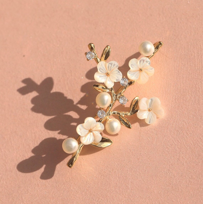May include: Gold tone brooch with a floral design featuring white flowers and pearls. The flowers are made of a pearlescent material.