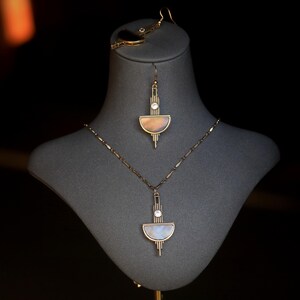 Pode incluir: Um conjunto de colar e brincos de ouro com um design geom&eacute;trico que apresenta uma forma de meia lua e um cristal transparente. A joia &eacute; exibida em um busto de manequim cinza.