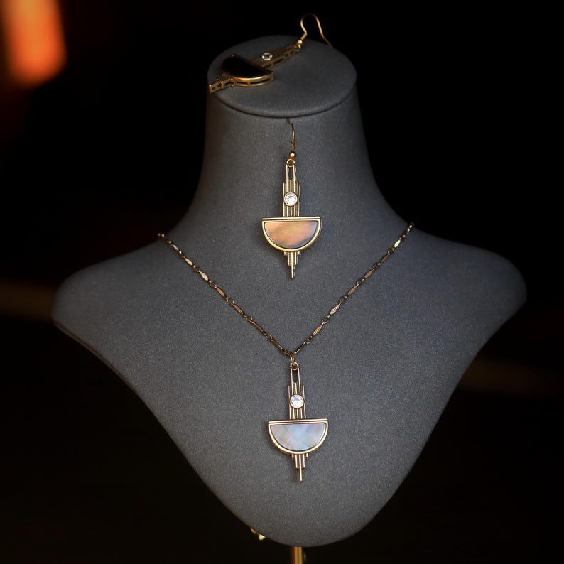 Pode incluir: Um conjunto de colar e brincos de ouro com um design geom&eacute;trico que apresenta uma forma de meia lua e um cristal transparente. A joia &eacute; exibida em um busto de manequim cinza.