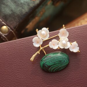 May include: A gold-toned brooch featuring a floral design with white blossoms and a green malachite stone. The brooch has a branch-like structure with delicate, pearl-centered flowers. The oval malachite stone has a distinctive green and black pattern.