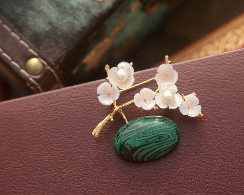 May include: A gold-toned brooch featuring a floral design with white blossoms and a green malachite stone. The brooch has a branch-like structure with delicate, pearl-centered flowers. The oval malachite stone has a distinctive green and black pattern.