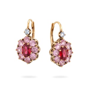 May include: A pair of gold earrings. Each earring has a central oval red gemstone, encircled by pink gemstones in a floral pattern. A small, round, clear gemstone is set above the floral design.