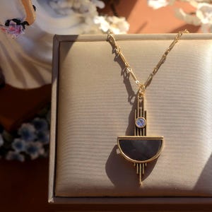 Pode incluir: Um colar de ouro com um pingente geom&eacute;trico que apresenta um design de semic&iacute;rculo preto e dourado e uma pequena pedra transparente no centro.
