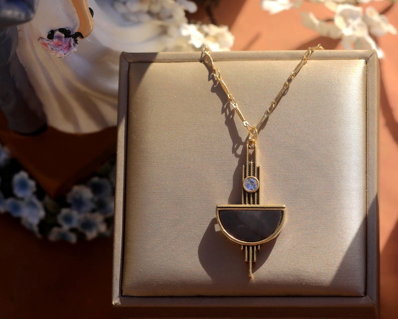 Pode incluir: Um colar de ouro com um pingente geom&eacute;trico que apresenta um design de semic&iacute;rculo preto e dourado e uma pequena pedra transparente no centro.