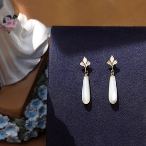 May include: A pair of white teardrop-shaped earrings with gold accents and a floral design. The earrings are on a dark blue background.