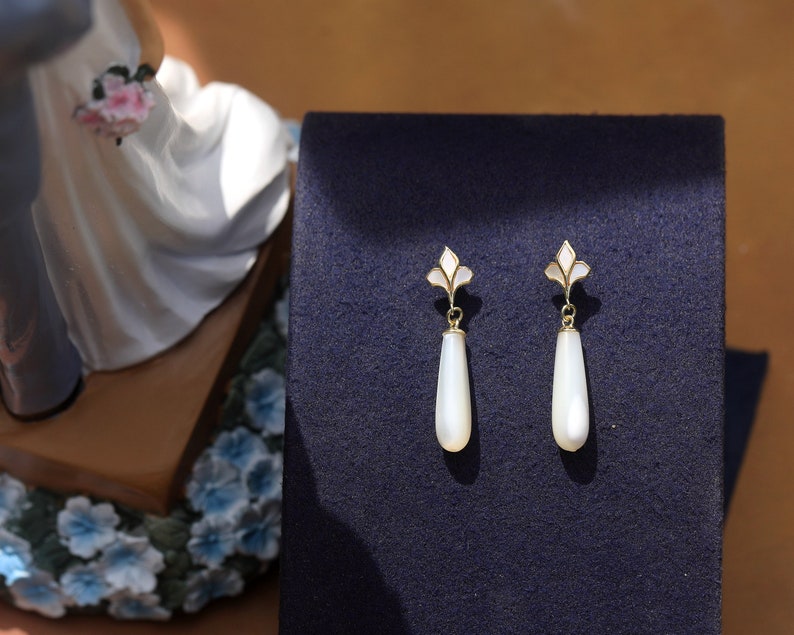May include: A pair of white teardrop-shaped earrings with gold accents and a floral design. The earrings are on a dark blue background.