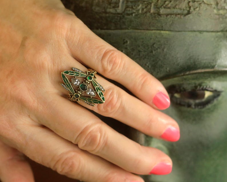 Pode incluir: Um anel de ouro com um design de lib&eacute;lula. A lib&eacute;lula tem asas de esmalte verde e strass transparentes e verdes.