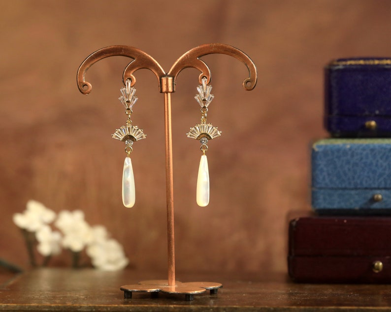 Op de afbeelding: Een paar gouden oorbellen met een waaiervormig ontwerp en een parelmoerachtige traanvormige hanger. De oorbellen worden getoond op een gouden standaard tegen een bruine achtergrond.