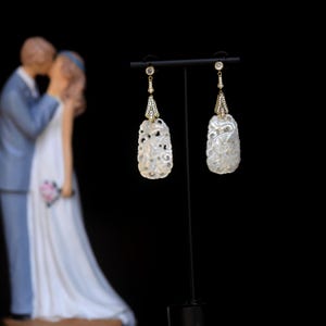 May include: A pair of white carved earrings with gold accents and clear stones hanging from a black stand. The earrings are in the shape of a teardrop with intricate floral designs.