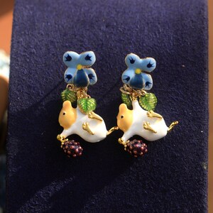 May include: A pair of gold-tone earrings featuring a white and yellow ceramic animal with a blue flower and green leaves hanging from it. The animal is holding a purple berry.