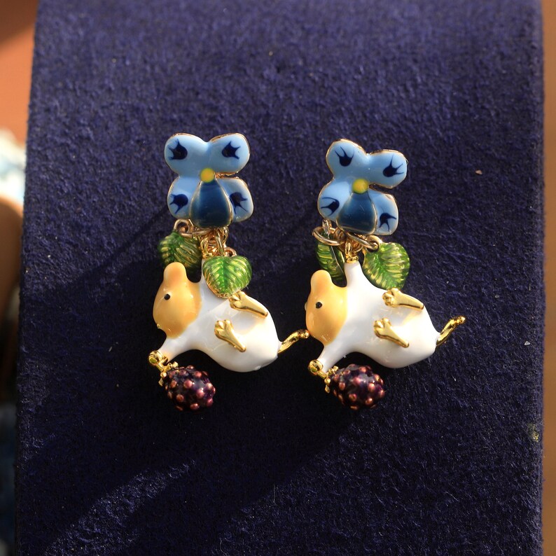 May include: A pair of gold-tone earrings featuring a white and yellow ceramic animal with a blue flower and green leaves hanging from it. The animal is holding a purple berry.