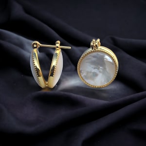 May include: A pair of gold hoop earrings with a white mother of pearl inlay. The earrings are on a black fabric background.