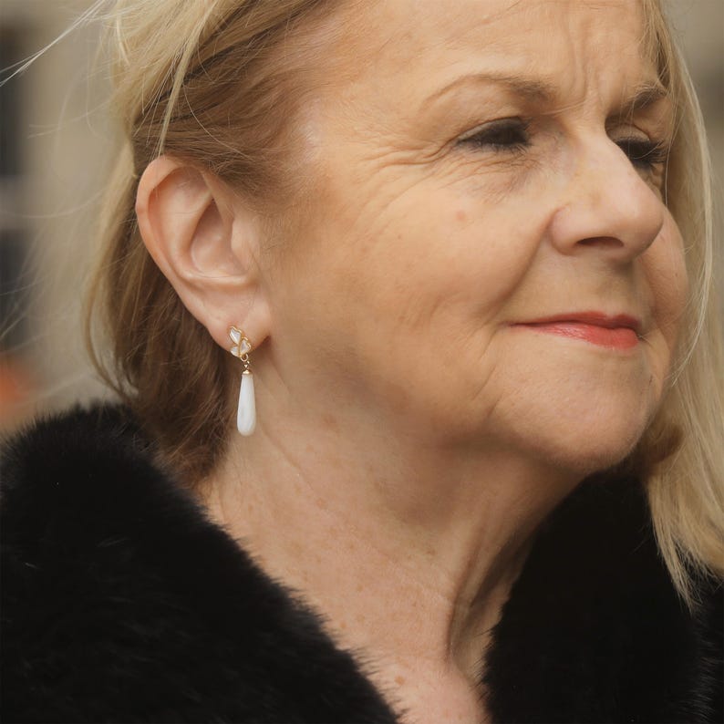 May include: A woman wearing a black fur coat with a pair of gold and white teardrop earrings.