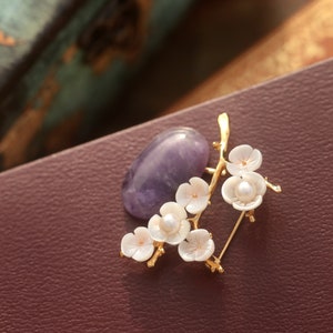 May include: A gold-toned brooch featuring a branch of white flower blossoms with pearl centers, paired with a purple gemstone. The brooch is set against a dark burgundy background, creating a contrast that highlights the delicate floral design.