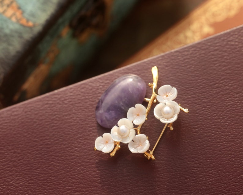 May include: A gold-toned brooch featuring a branch of white flower blossoms with pearl centers, paired with a purple gemstone. The brooch is set against a dark burgundy background, creating a contrast that highlights the delicate floral design.