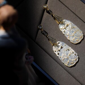 May include: A pair of white mother of pearl earrings with intricate carvings of birds and floral designs. The earrings are accented with black gemstones and have a gold tone finish.