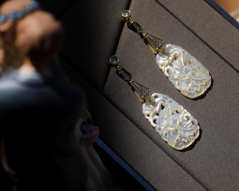 May include: A pair of white mother of pearl earrings with intricate carvings of birds and floral designs. The earrings are accented with black gemstones and have a gold tone finish.