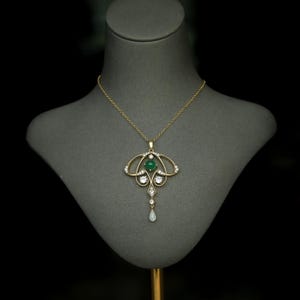 May include: A gold-toned pendant necklace with an Art Nouveau design. The necklace features a central green gemstone, surrounded by smaller clear stones and a teardrop-shaped pearl. The necklace is displayed on a gray mannequin.