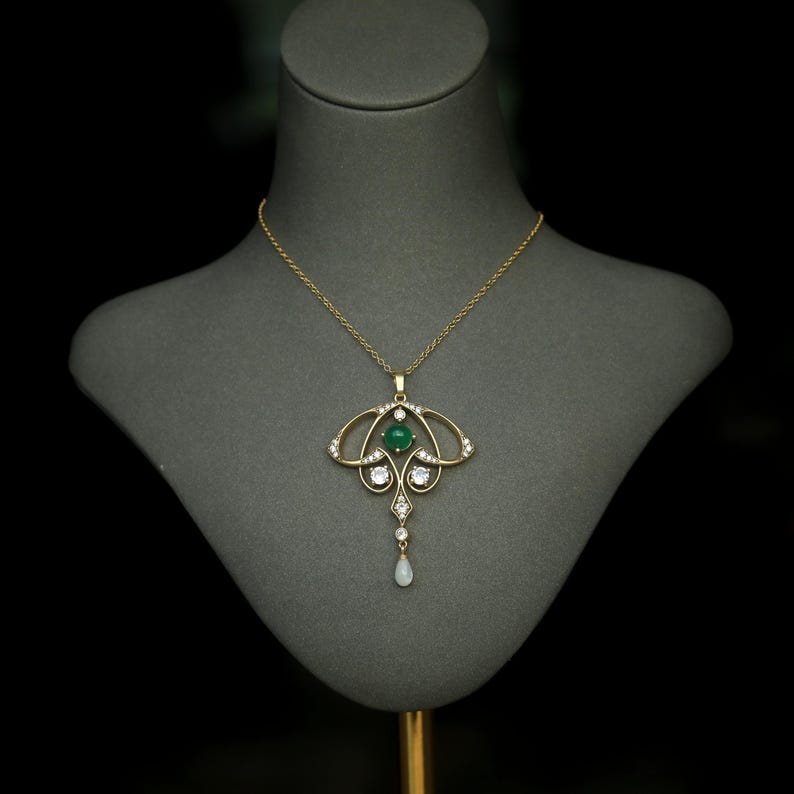 May include: A gold-toned pendant necklace with an Art Nouveau design. The necklace features a central green gemstone, surrounded by smaller clear stones and a teardrop-shaped pearl. The necklace is displayed on a gray mannequin.