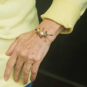 May include: A gold bracelet with a delicate design featuring a pearl and three small birds with green and yellow enamel. The bracelet is on a person's wrist.