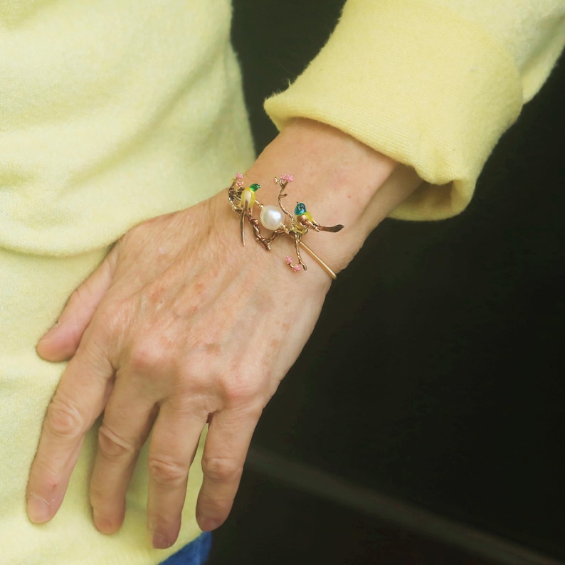 May include: A gold bracelet with a delicate design featuring a pearl and three small birds with green and yellow enamel. The bracelet is on a person's wrist.