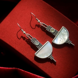 May include: A pair of silver-toned drop earrings featuring a geometric design with vertical lines, a small circular clear stone, and a semi-circular white iridescent inlay. The earrings are attached to standard silver-toned metal fishhook wires.