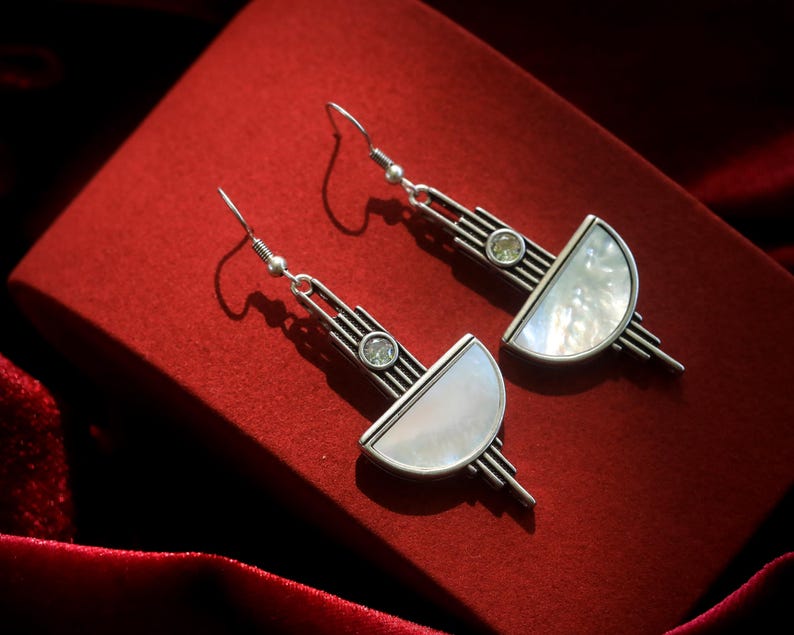 May include: A pair of silver-toned drop earrings featuring a geometric design with vertical lines, a small circular clear stone, and a semi-circular white iridescent inlay. The earrings are attached to standard silver-toned metal fishhook wires.