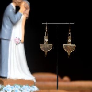 Op de afbeelding: Een paar gouden oorbellen met een halve maanvormige schelp en een klein strass-accent. De oorbellen hangen aan een zwarte standaard.