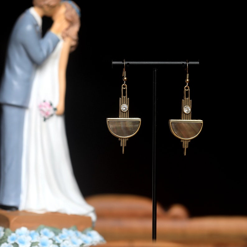 Op de afbeelding: Een paar gouden oorbellen met een halve maanvormige schelp en een klein strass-accent. De oorbellen hangen aan een zwarte standaard.