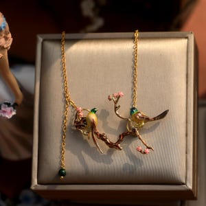 May include: A gold-colored necklace featuring two birds perched on a branch with pink blossoms. The birds have yellow, green, and brown details. A green bead hangs from the chain.