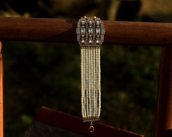 May include: A white pearl bracelet with a gold and clear rhinestone clasp. The bracelet is hanging from a wooden surface.