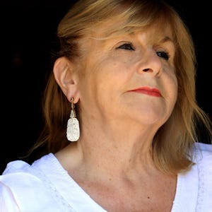 May include: A woman with blonde hair wearing a white shirt and dangling earrings with a white, textured design.