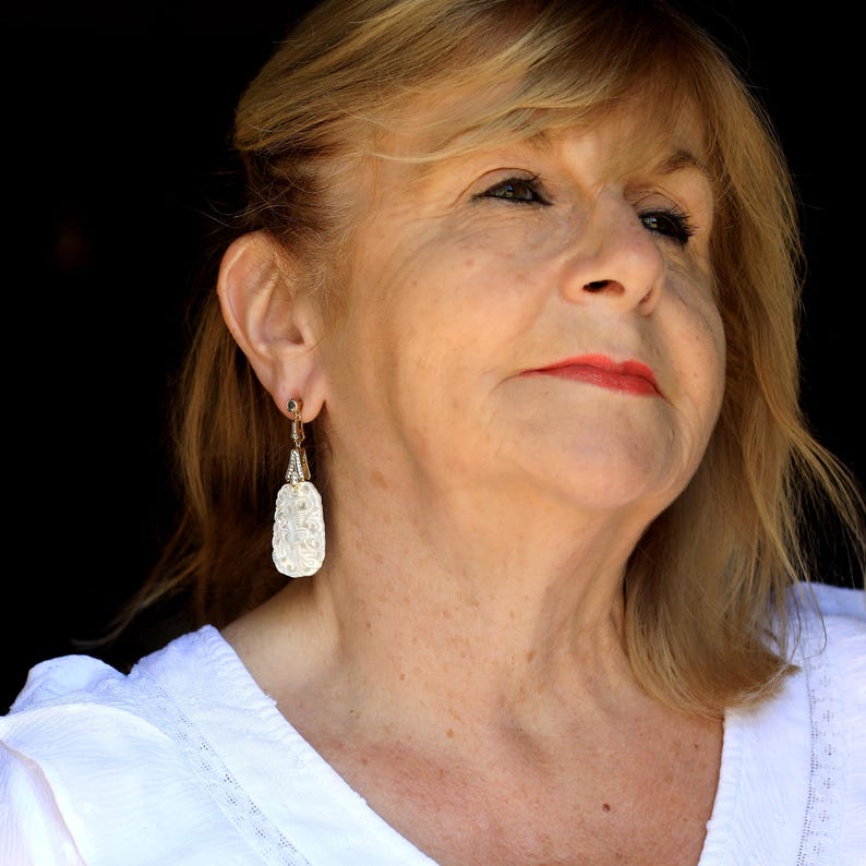May include: A woman with blonde hair wearing a white shirt and dangling earrings with a white, textured design.