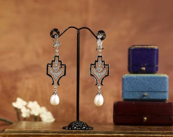 May include: A pair of silver dangle earrings with a geometric design and a pearl drop. The earrings are displayed on a black stand against a brown background.